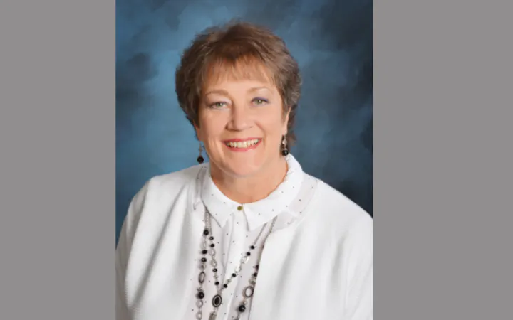 Portrait of a smiling woman in a white blazer with a layered necklace, against a blue backdrop.