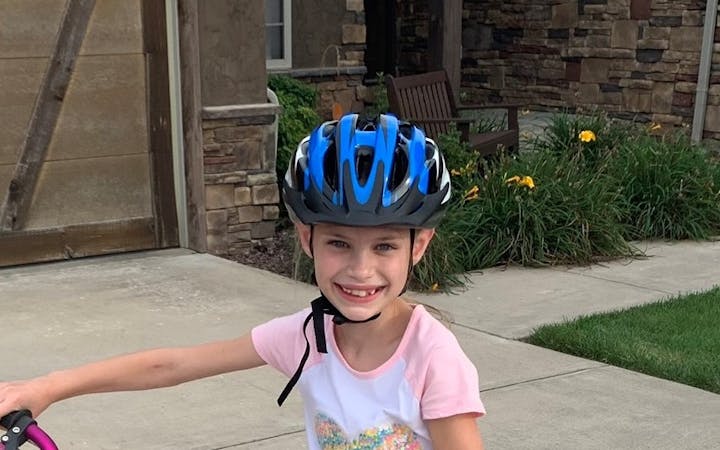 A young girl wearing a blue bicycle helmet and standing on her pink bike smiles into the camera