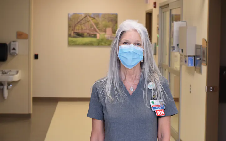 Nurse in gray scrubs wearing a blue mask in a hospital hallway, badge with RN visible.