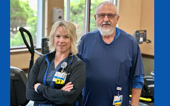 Two healthcare workers in scrubs stand in a rehab gym with exercise equipment.