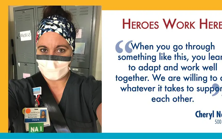 Nurse in scrubs and mask beside a 'Heroes Work Here' poster displaying the name Cheryl Nold.