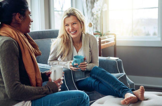 Two women sit together in a cozy living room, talking and drinking coffee