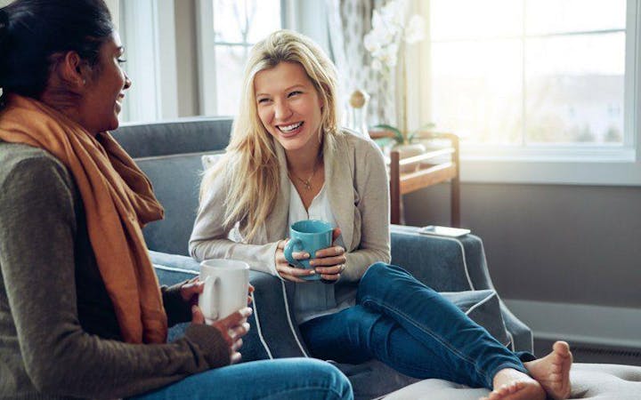 Two women sit together in a cozy living room, talking and drinking coffee