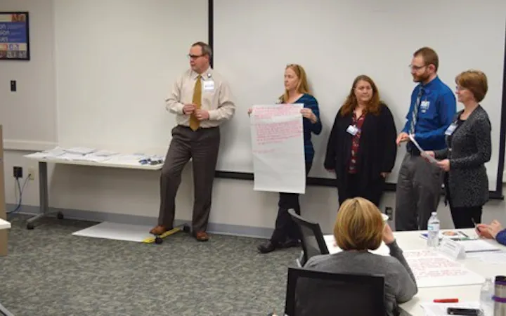 Six people stand at the front of a meeting room; several hold sheets of paper.