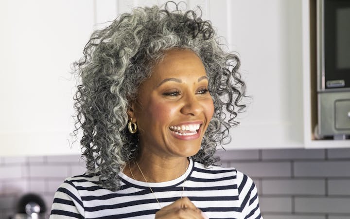 A middle-aged woman holding a healthy green drink stands in her kitchen and smiles.