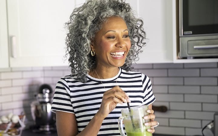 A middle-aged black woman with curly gray hair stands in her kitchen and smiles, holding a healthy green smoothie.