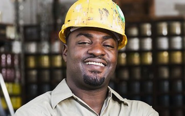 A worker in a hard hat stands with his hands folded and smiles into the camera