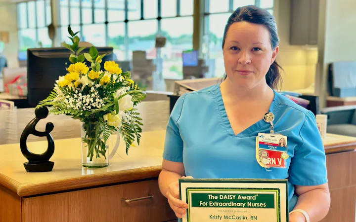 Kristy McCaslin, RN, nurse in blue scrubs with DAISY Award certificate beside flowers.
